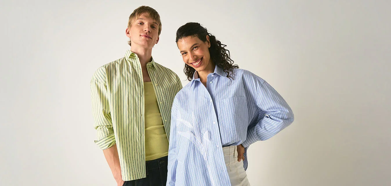 Dos personas están de pie sonriendo frente a un fondo liso, vistiendo camisas de rayas holgadas y pantalones casuales, posando con confianza.