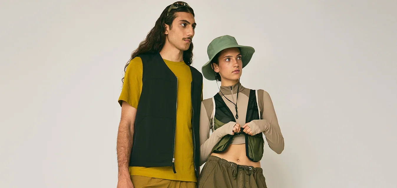 Dos personas de pie frente a un fondo liso, vestidas con ropa casual en tonos terrosos, con chalecos y pantalones o pantalones cortos relajados.