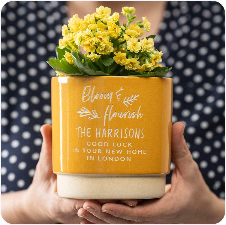 A personalised yellow plant pot engraved with a family name and address, held against a dark background with yellow flowers