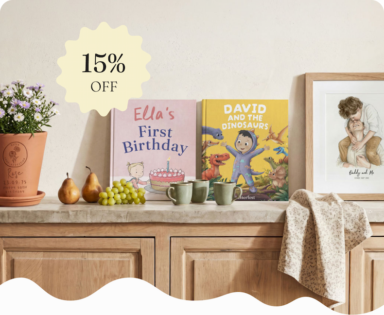 A personalised first birthday story book, a personalised dinosaur book and a framed watercolour portrait displayed on a wooden sideboard with a plant and fruit, with a 15% off badge overlay