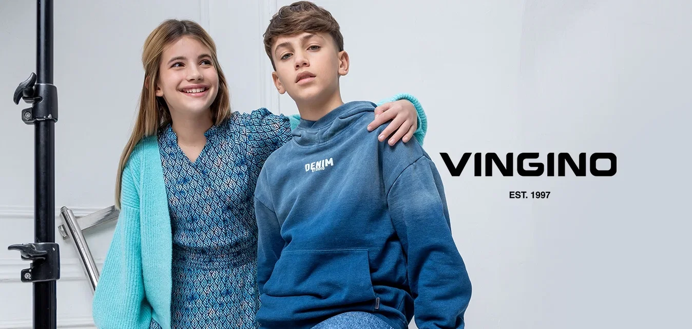 Twee kinderen poseren in een studio. Eén draagt een blauwe jurk met patroon en een lichtblauwe cardigan, terwijl de ander een blauwe hoodie met verloopkleur en een spijkerbroek draagt.
