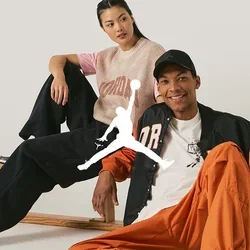 Twee personen zitten casual op een houten bank. De man draagt een oranje en zwarte jas, een wit T-shirt, oranje broek en grijze sneakers. De vrouw heeft een roze en beige gebreid topje aan met zwarte broek en zwarte sneakers.