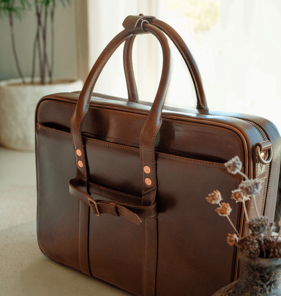 Close-up of a brown leather briefcase with rolled top handles and copper rivets on a table near dried flowers.