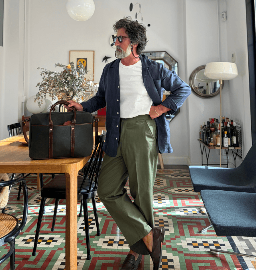 A man in a navy linen shirt and olive trousers stands by a wood table holding a black leather briefcase.