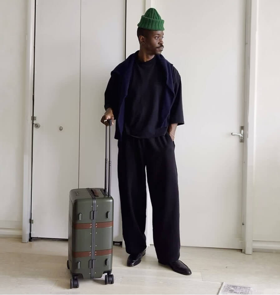 A man in black loungewear and a green beanie stands with an olive green hardshell suitcase in a white hallway.