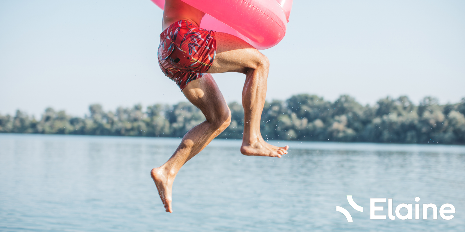 Mann springt mit pinkem Schwimmring ins Wasser, trägt kurze Badehose, sonniger Tag am See.