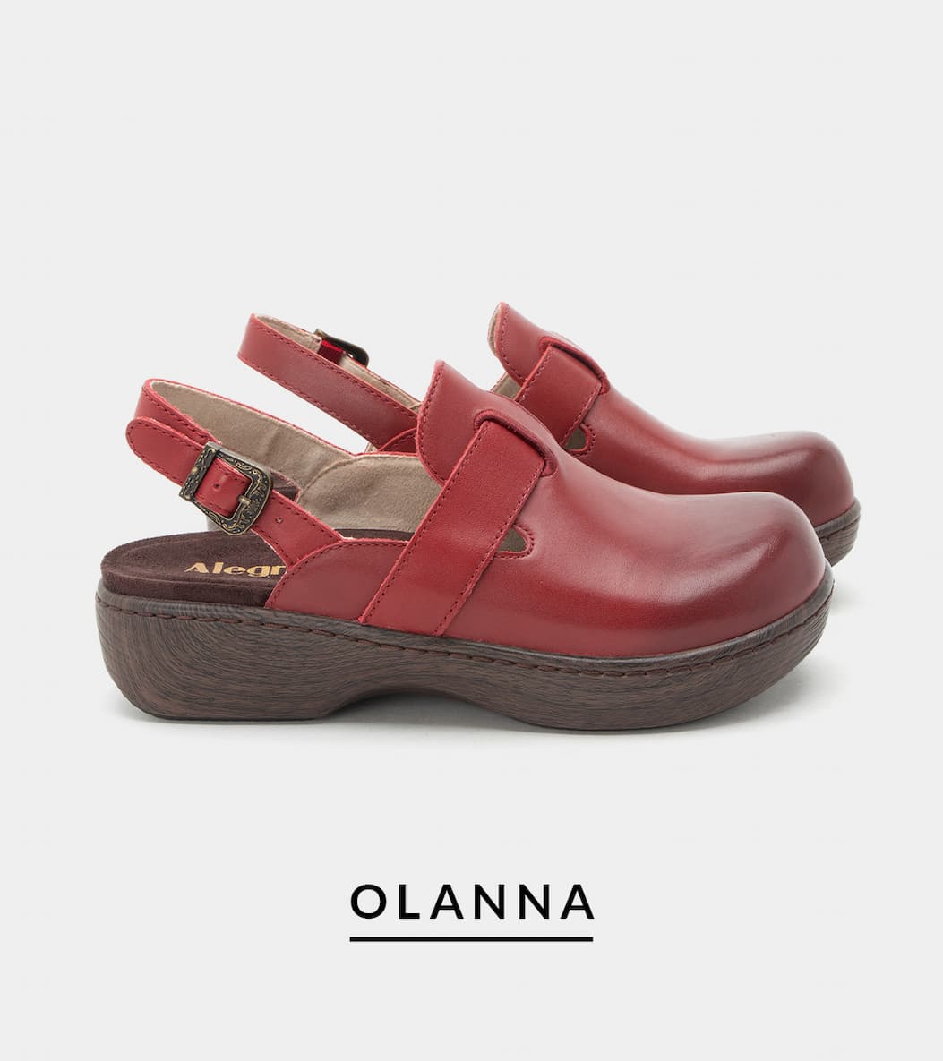 A side profile view of a pair of "Olanna" red leather closed-toe clogs. The shoes feature a slingback design with an adjustable strap and an antique brass buckle, set on a dark wood-grain look rocker outsole.