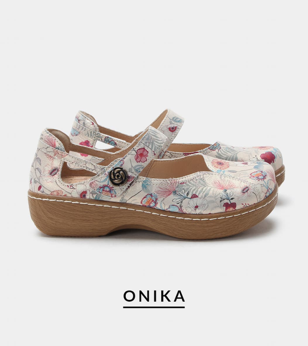 A side profile view of a pair of "Onika" Mary Jane shoes. The shoes feature an off-white upper with a soft watercolor floral and leaf print pattern, cut-out side detailing, an adjustable strap with a dark metal rose-shaped button, and a light-colored wood-grain look rocker outsole.