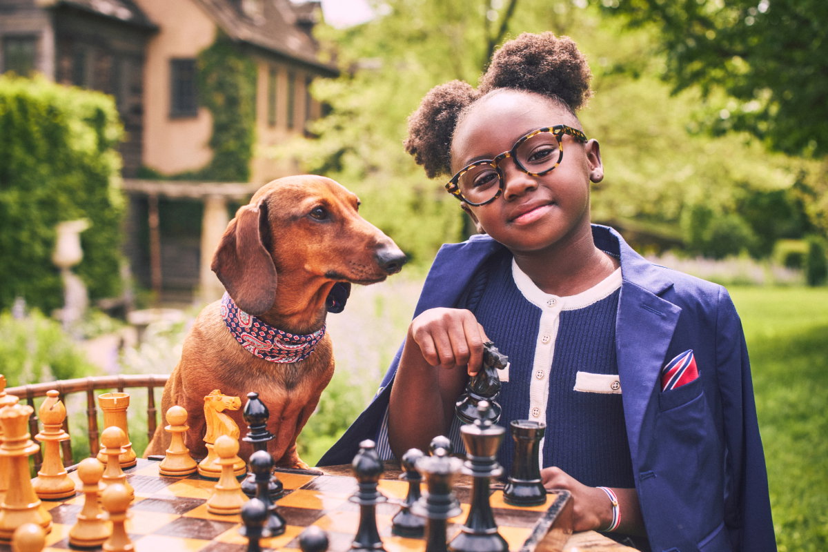 A young girl wearing glasses and a navy blazer playing chess outdoors with a dachshund wearing a bandana