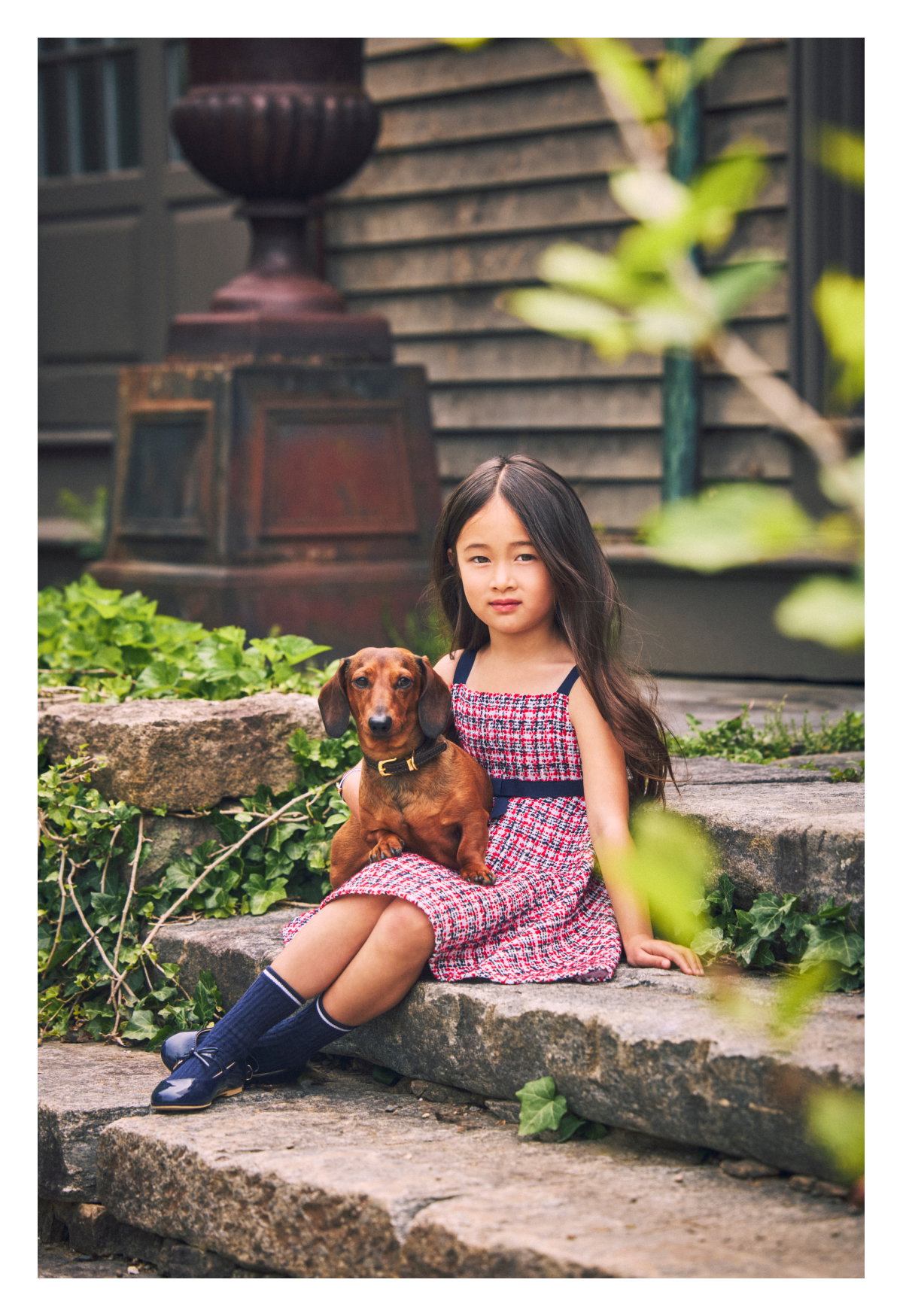 A young girl in a red tweed dress sitting on stone steps holding a dachshund puppy