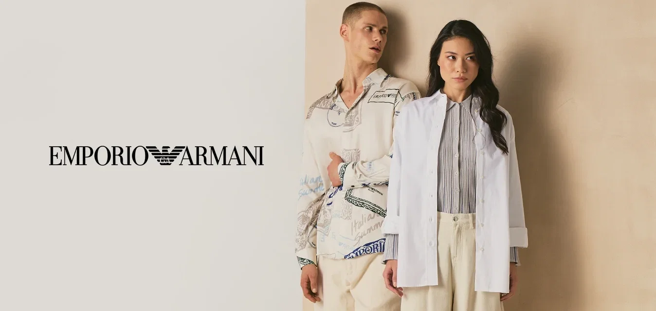 Due persone sono appoggiate a un pannello beige, una indossa una camicia a motivo e pantaloni color crema, l'altra un blazer bianco e pantaloni larghi color crema.
