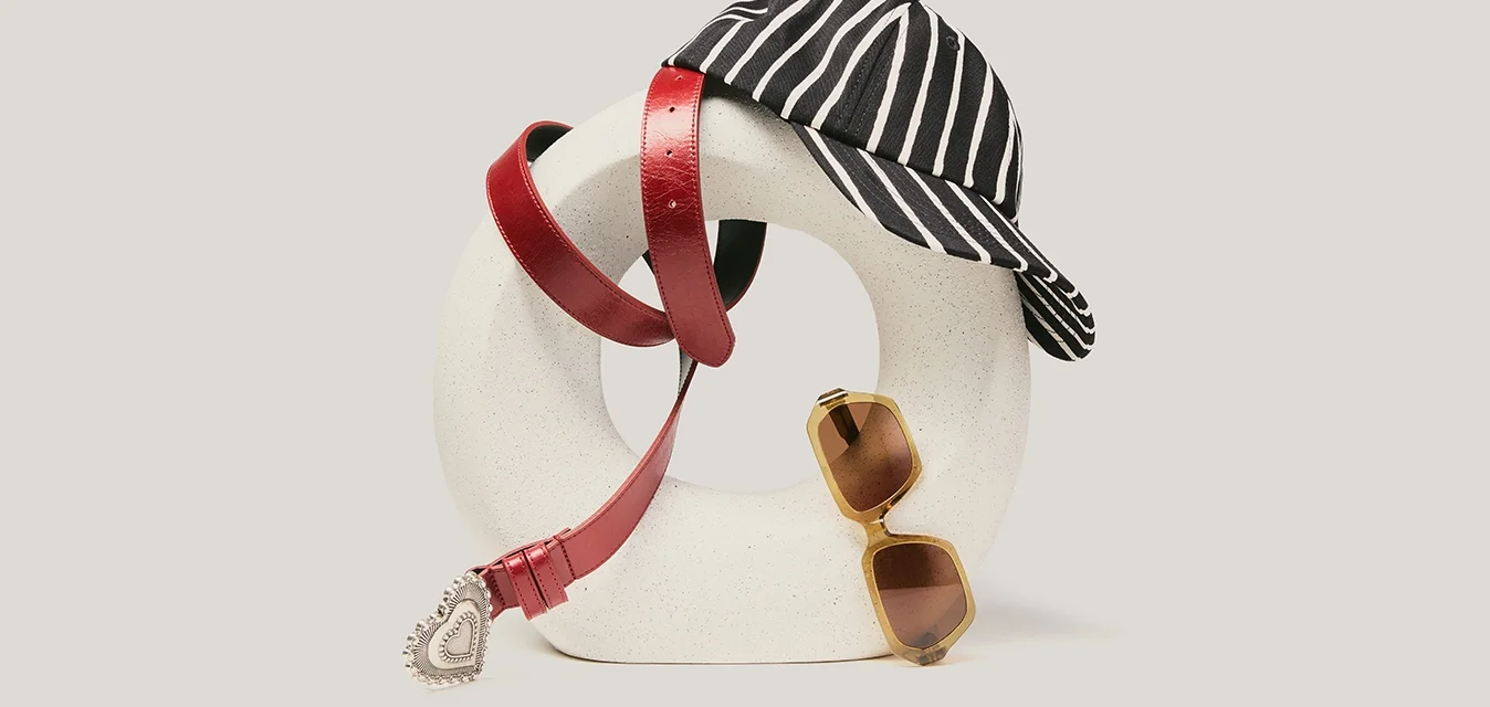 Scultura astratta bianca con una cintura rossa drappeggiata sopra, un berretto a righe bianco e nero sulla sommità e occhiali da sole con lenti marroni appoggiati contro di essa.