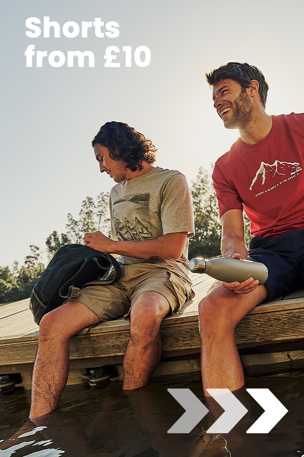 Two people sit on a wooden dock with their feet in the water, wearing casual outdoor shorts, promoting Peter Storm shorts from £10.