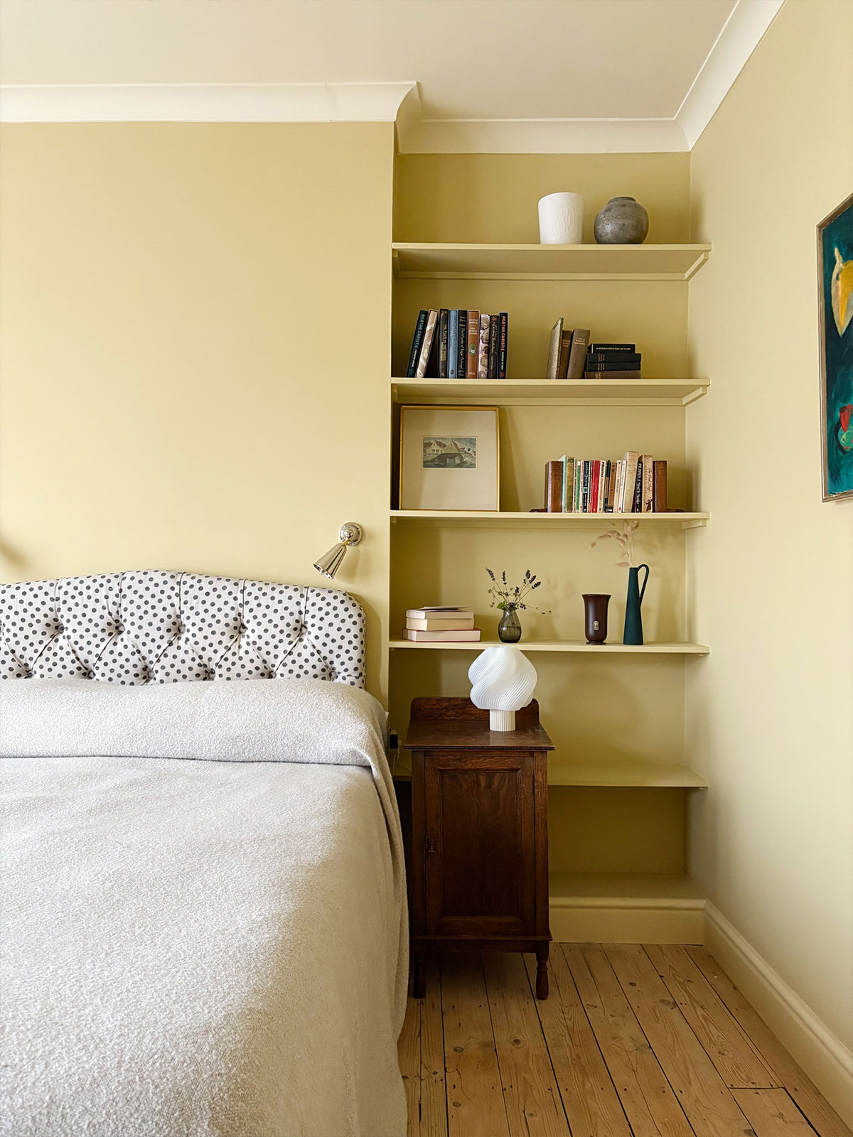 Soft yellow bedroom with built-in shelves, a spotted headboard, and a wooden bedside table.
