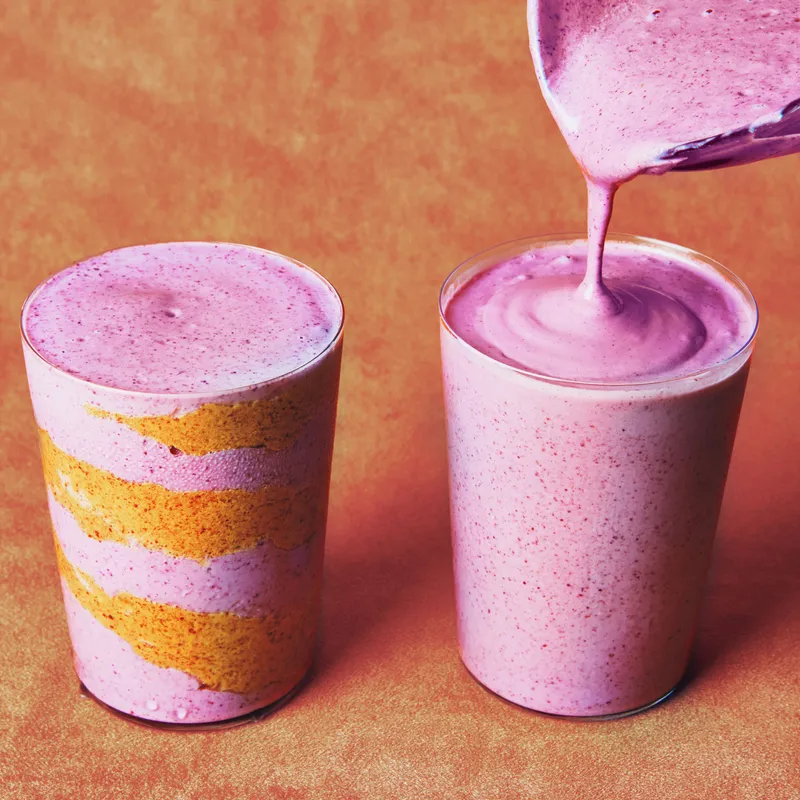 Blended mixed berries, cottage cheese, water, and almond butter being poured into two glass cups.