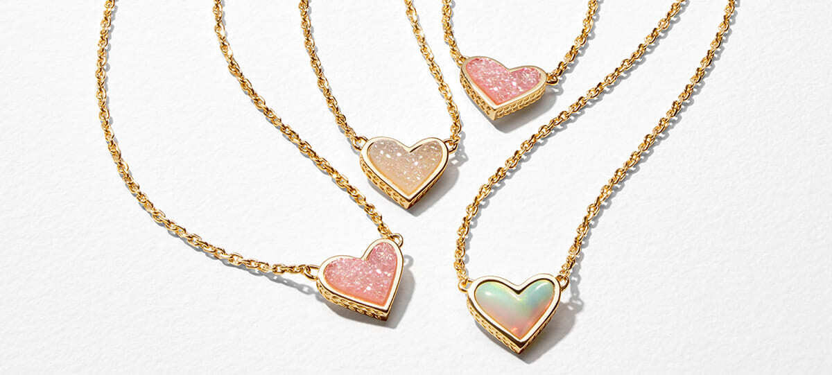 Four gold heart pendant necklaces featuring pastel stone inlays, arranged on a light textured background.