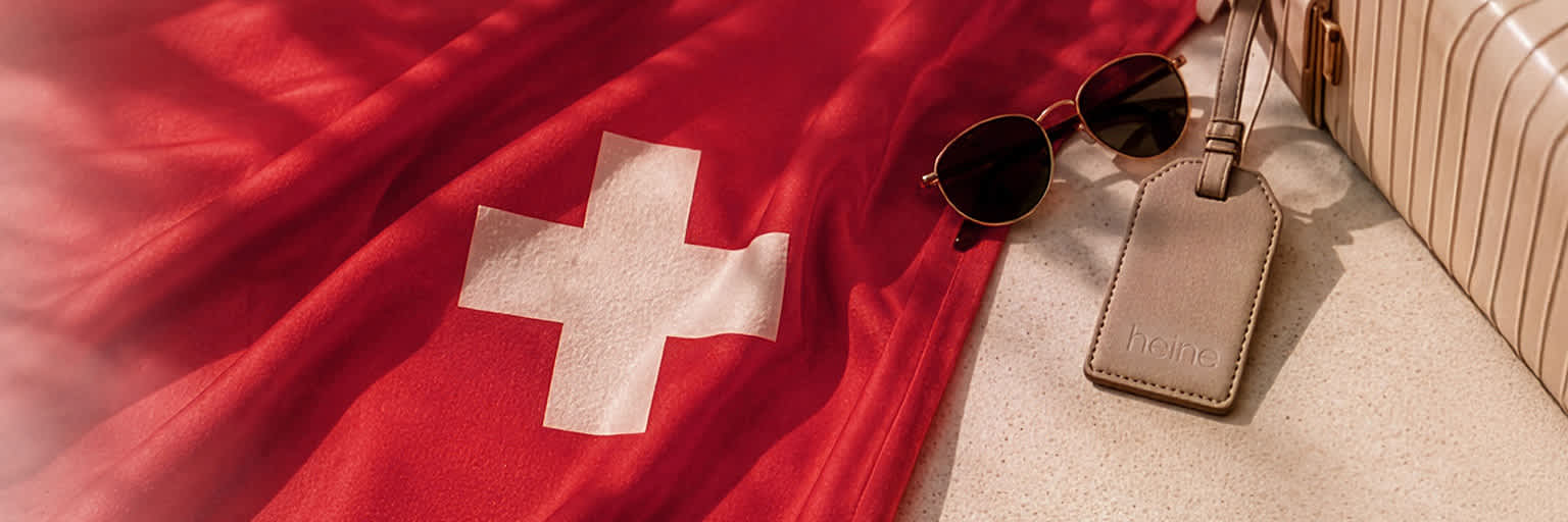 Eine Werbeanzeige mit dem Text Auf dem Bild ist kein Text zu sehen. Der Hintergrund zeigt eine ausgebreitete Schweizer Flagge, daneben liegen eine Sonnenbrille, ein beiger Koffer und ein Kofferanhänger mit der Prägung 'heine' auf hellem Untergrund. 