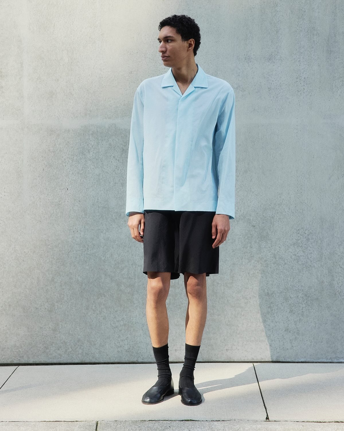 Young man wearing a light-blue long-sleeve shirt, black shorts, black socks and shoes standing against a concrete wall