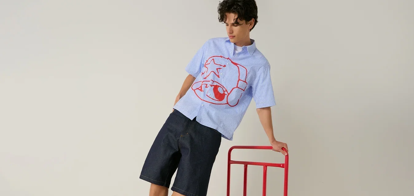 Jonge man die balanceert op een rode platformwagen, draagt een blauw shirt met rode print, zwarte korte broek, witte sokken en rode sneakers, tegen een effen achtergrond.