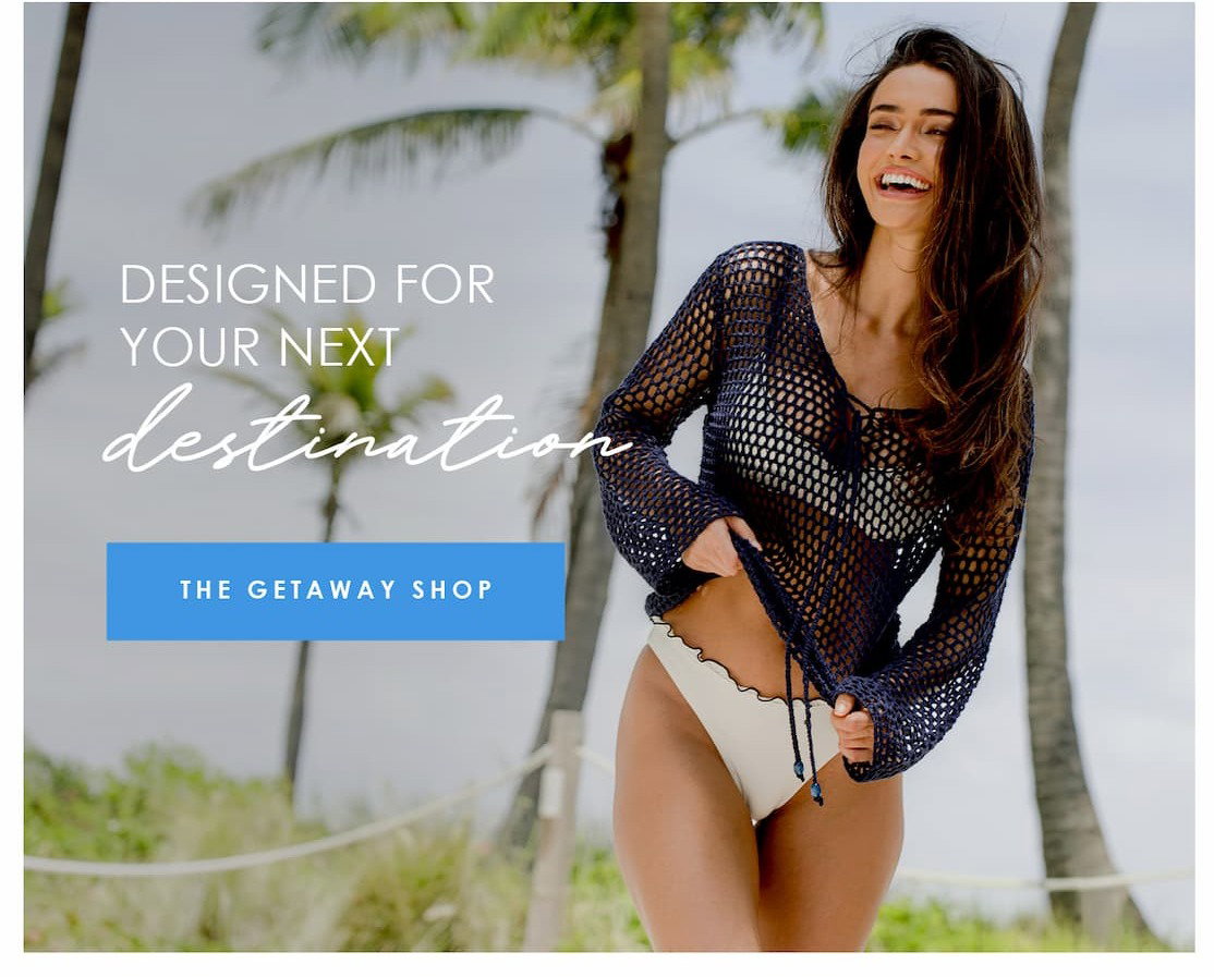 Smiling model in a navy crochet cover-up and white swim bottoms at a tropical destination, with text reading “Designed for your next destination” and a button for The Getaway Shop.