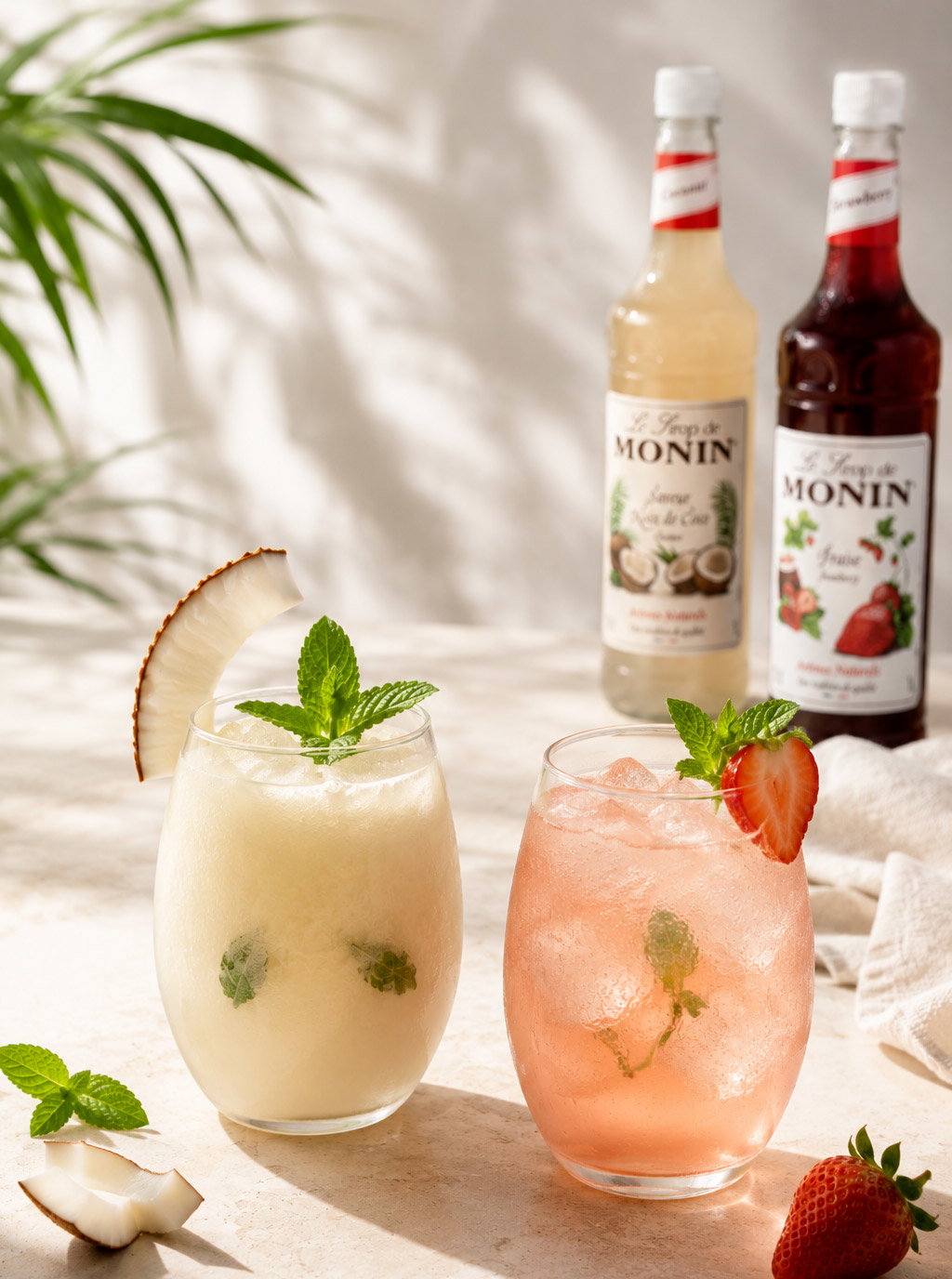 Frozen coconut cocktail and pink strawberry iced mocktail garnished with mint and berry; Monin syrup bottles background.