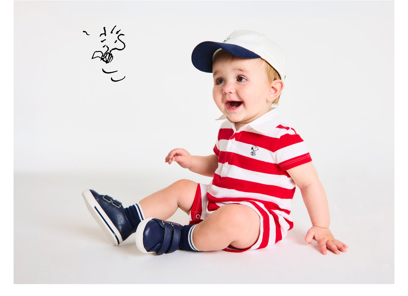 Adorable smiling baby in red and white striped Snoopy romper, navy cap, and navy sneakers sitting on white background with cartoon doodle