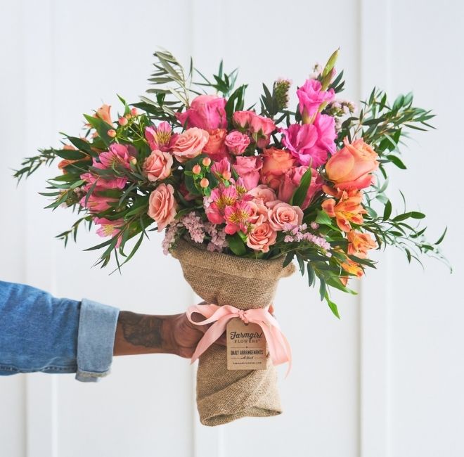 A hand holds up a small burlap-wrapped Fun Size bouquet of coral and pink roses with orange accents and greenery, tied with a blush ribbon.