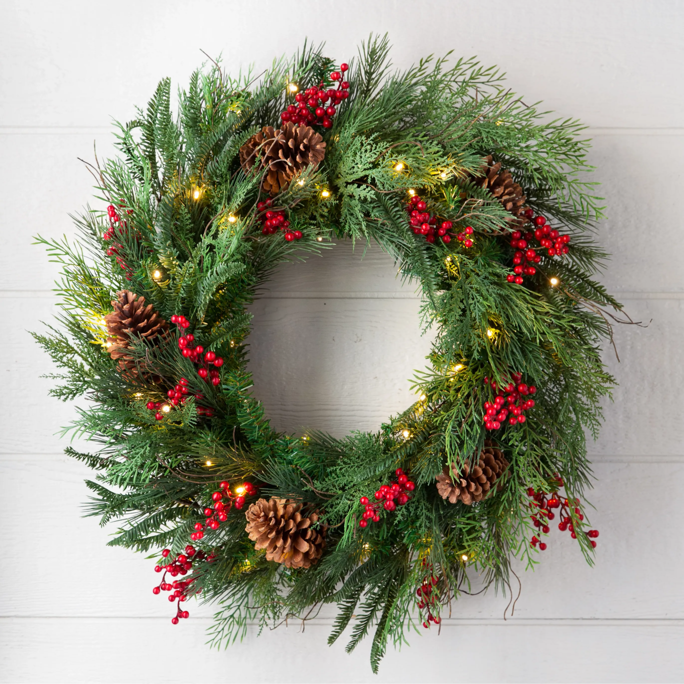 Pre-lit Christmas wreath with pinecones, red berries, and mixed greenery on a white wall.