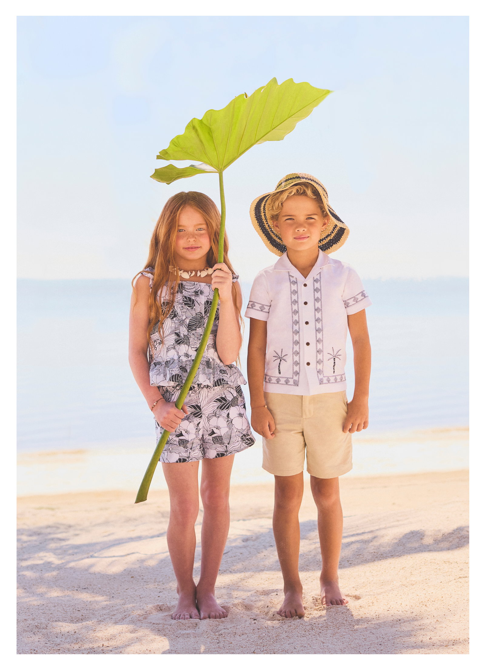 Two children standing barefoot on a sandy beach, one holding a large tropical leaf, wearing summer resort-style clothing