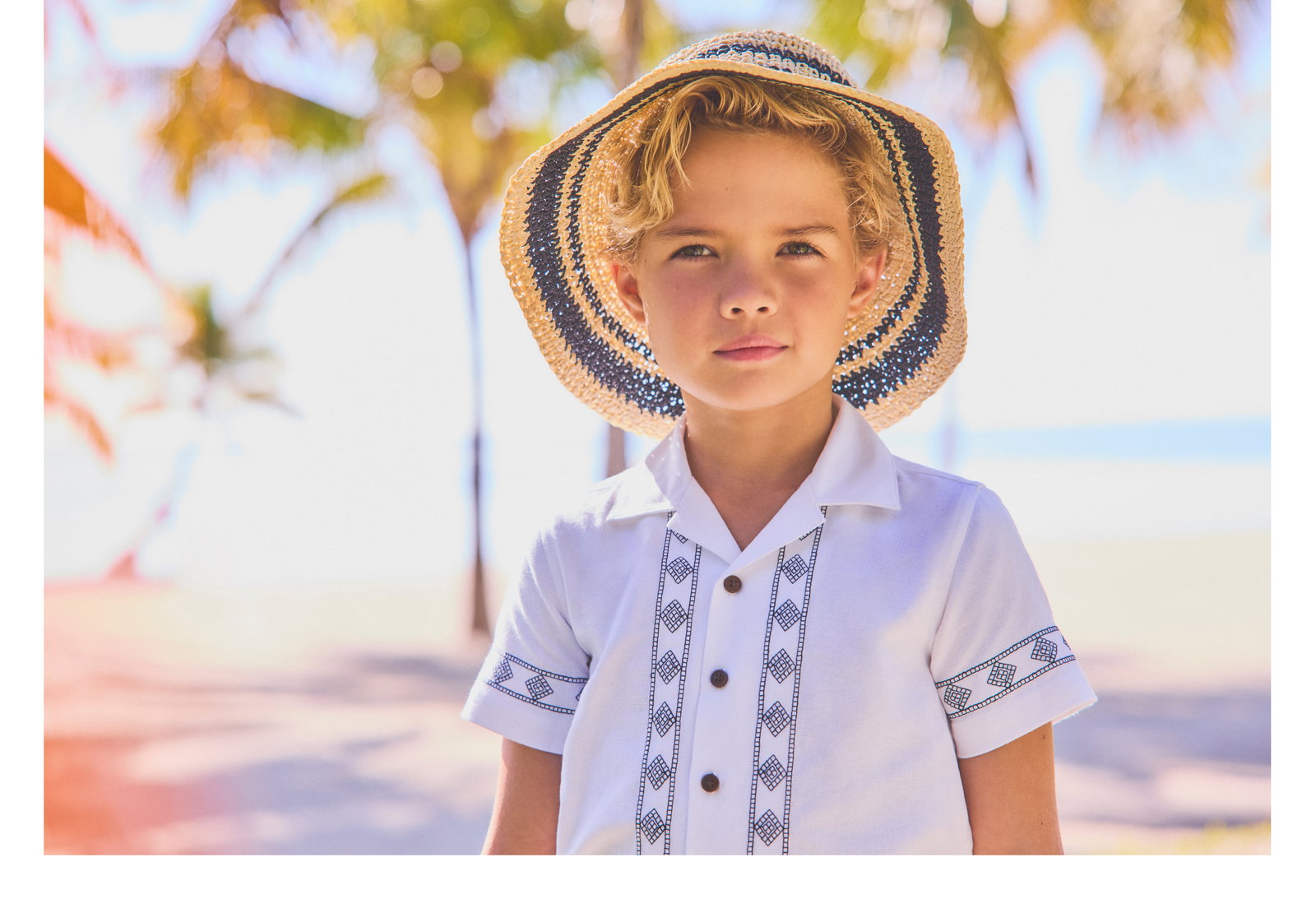 Young boy wearing a straw hat with navy stripes and a white embroidered guayabera shirt, standing outdoors near palm trees