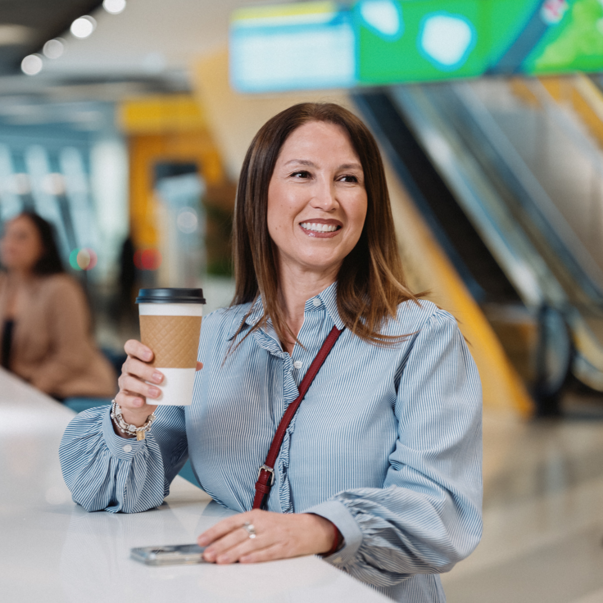 A woman sits at a bright counter holding a takeaway coffee cup and smiling. She is in a contemporary indoor space with escalators and colorful architectural elements in the background. The setting appears to be a public or commercial building, such as an office lobby or mall. A smartphone rests on the counter near her hand, adding a casual, everyday feel to the scene.