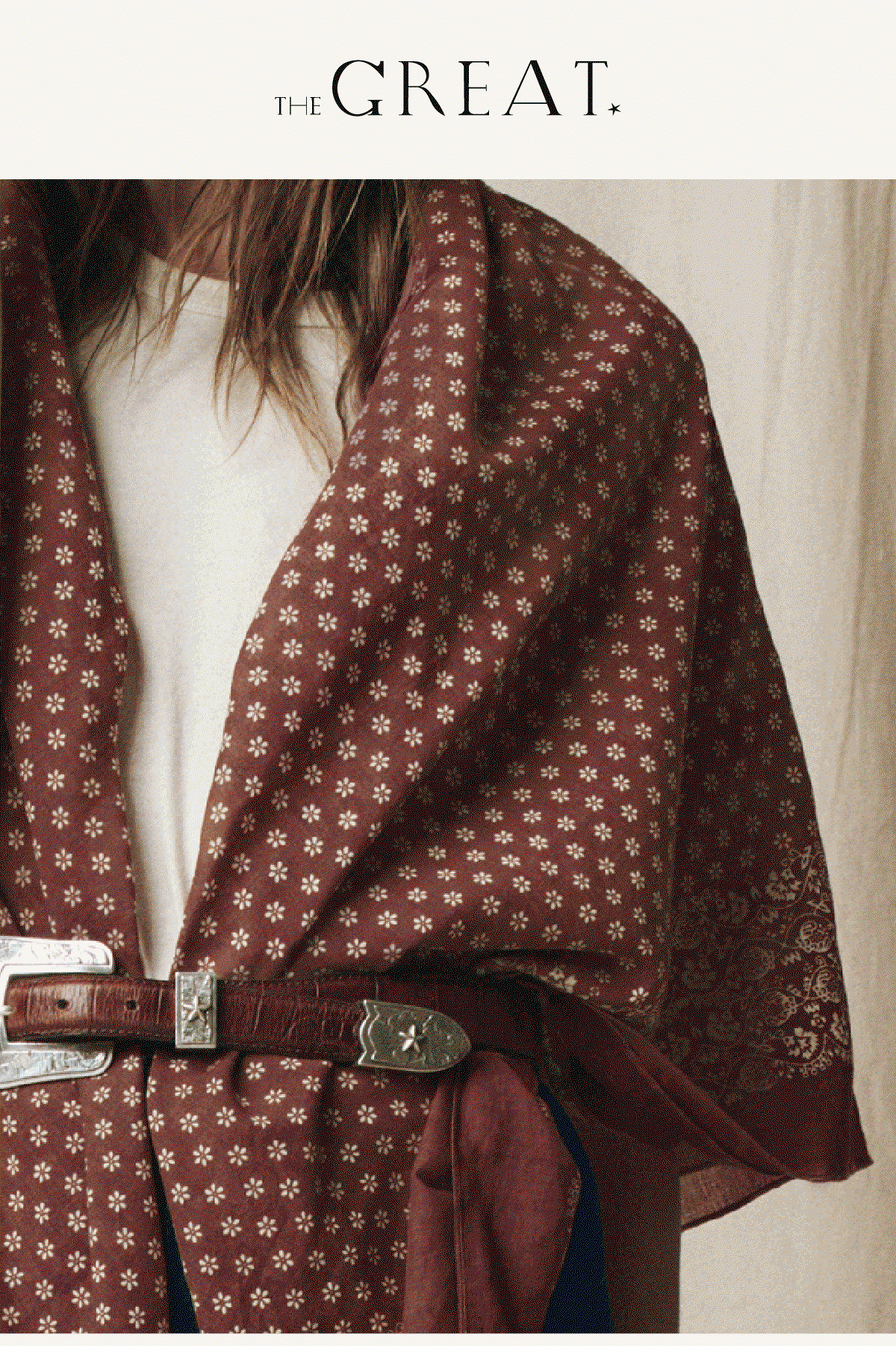 Model wearing an oversized rust bandana with a delicate vintage-inspired floral print, draped over a white tee and cinched at the waist with a brown leather belt featuring silver western hardware. The bandana's paisley border peeks through at the edge, highlighting its versatile oversized shape.
