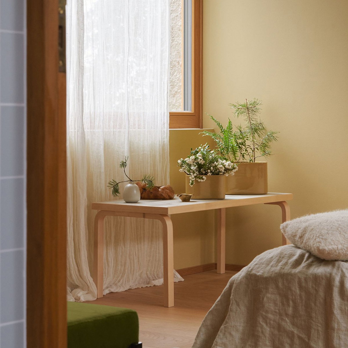 Panca Artek in betulla davanti alla finestra della camera da letto, con vasi di fiori e oggetti decorativi