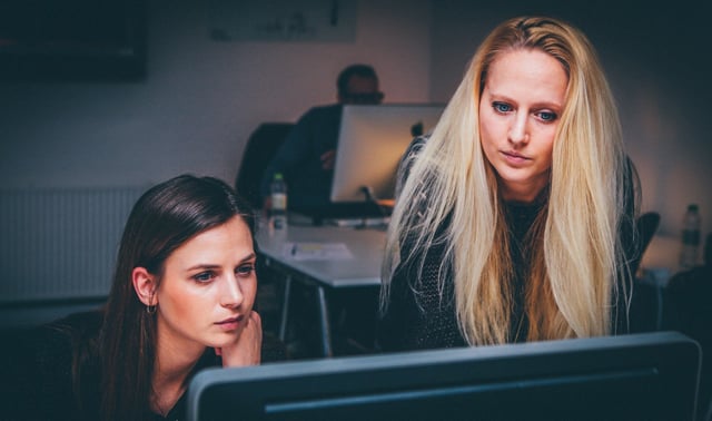 Two women focus attentively on a computer screen in an office setting, with a person working in the background.