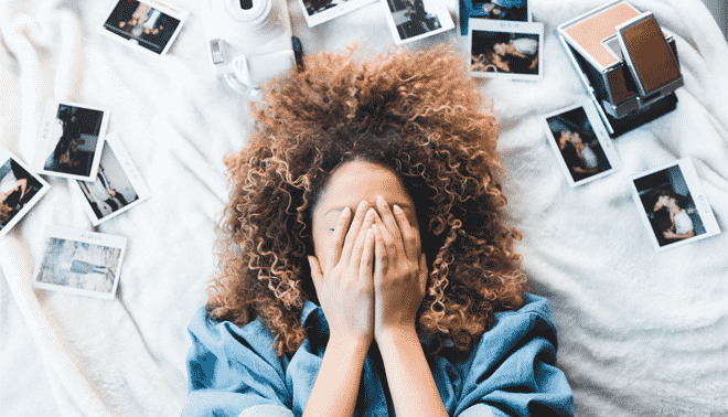 A person with curly hair lies on a bed with hands covering their face, surrounded by scattered Polaroid photos and a Polaroid camera.