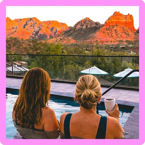 Image description: Two women in a pool with drinks looking out at red stone cliffs