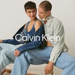 Twee jonge volwassenen, met blote voeten en in denim gekleed, zitten dicht naast elkaar op een houten bank tegen een effen achtergrond.