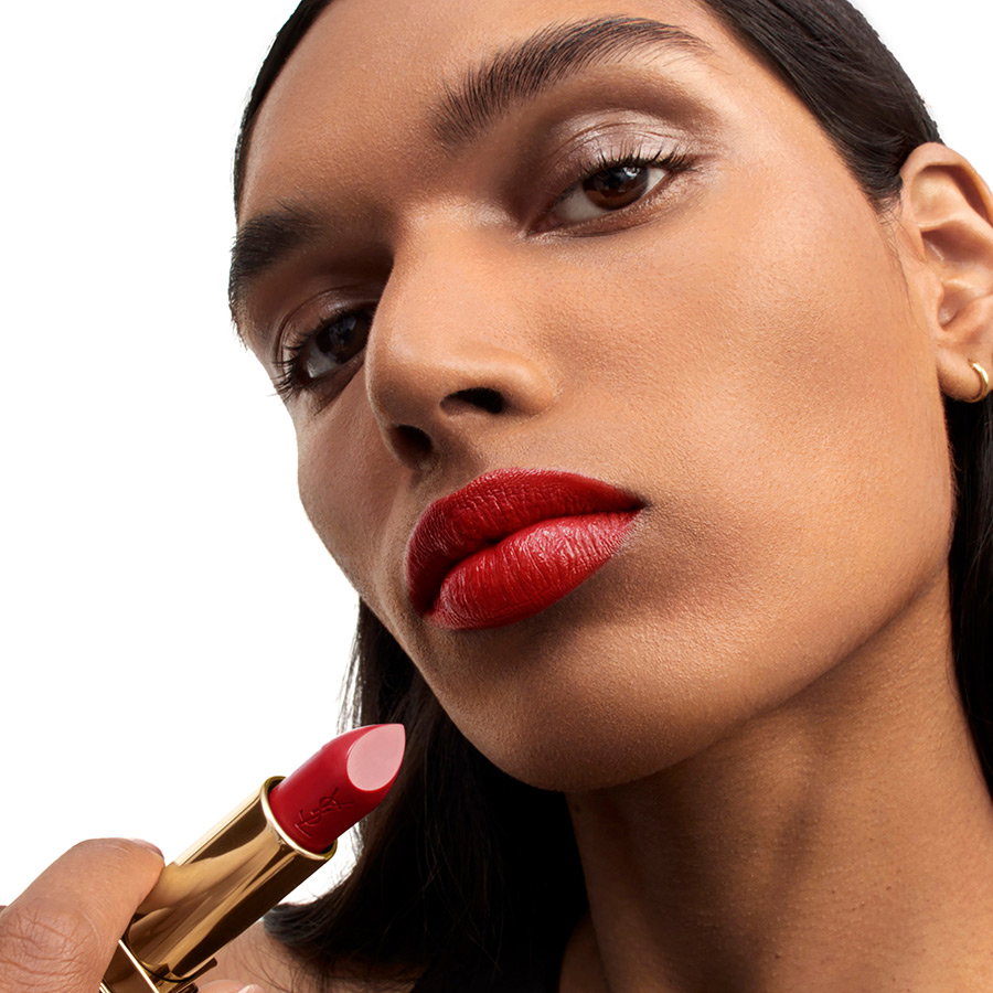 Close-up of a woman with warm medium-dark skin holding a bold red YSL lipstick to her perfectly painted lips.