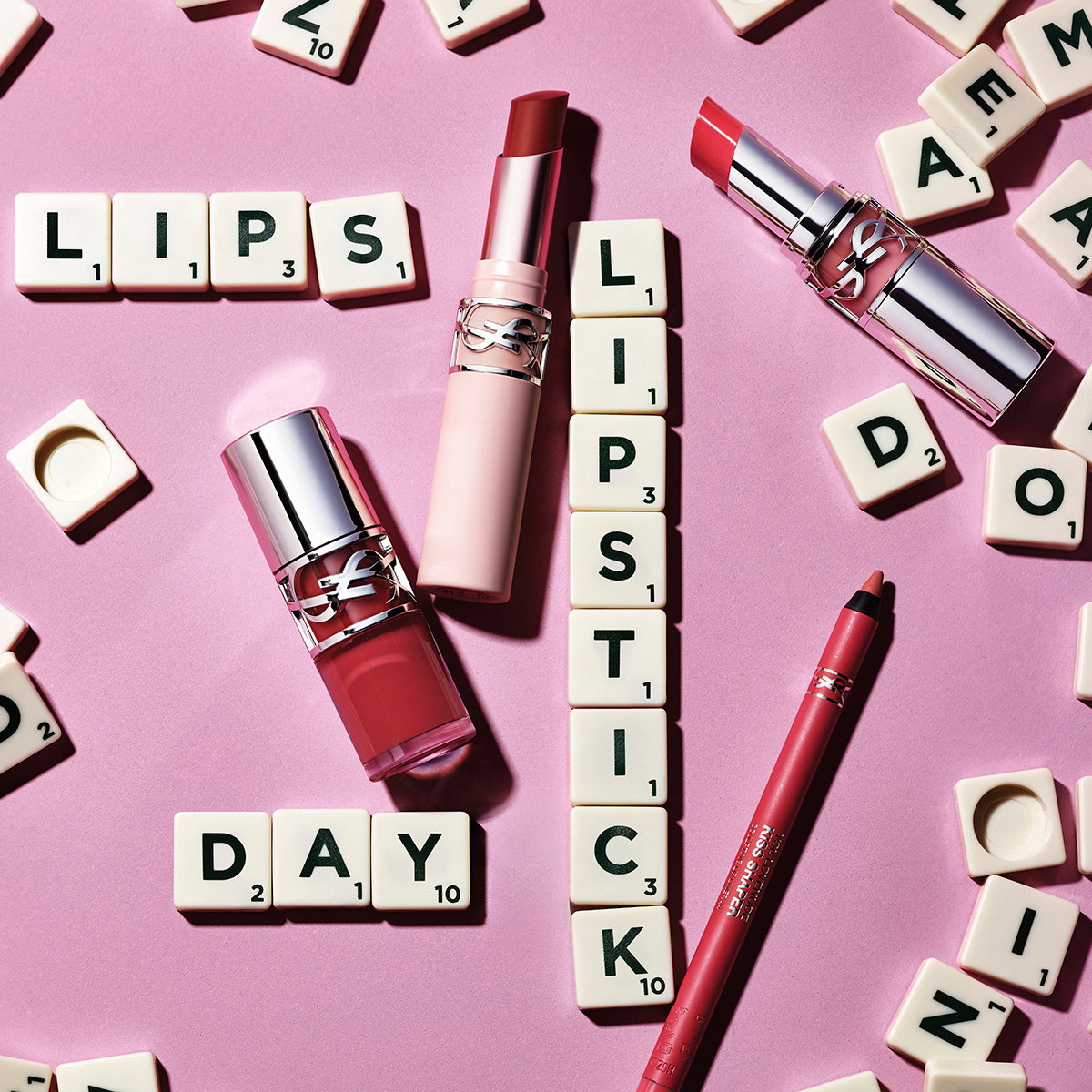 A selection of YSL lipsticks and a lip liner arranged on a pink surface with Scrabble tiles spelling "LIPS", "LIPSTICK", and "DAY".