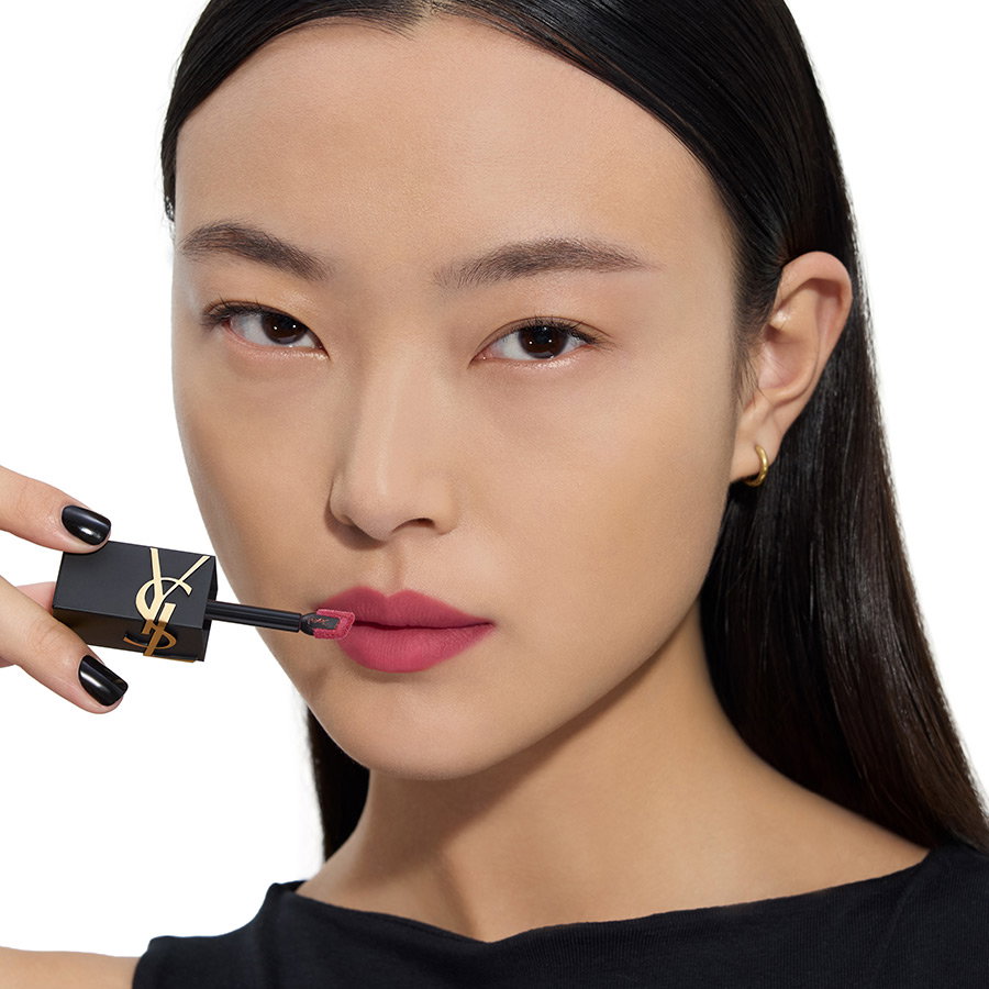 Close-up of an Asian woman with black nail polish applying a matte berry liquid lipstick using a YSL applicator.