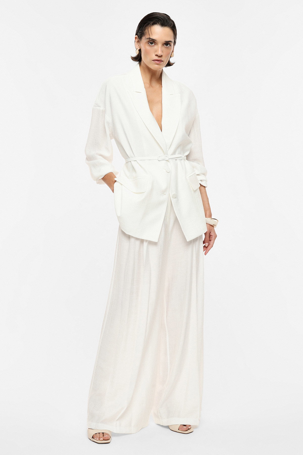 Woman in white belted blazer and wide-leg trousers standing in minimalist studio, hands in pockets, neutral expression
