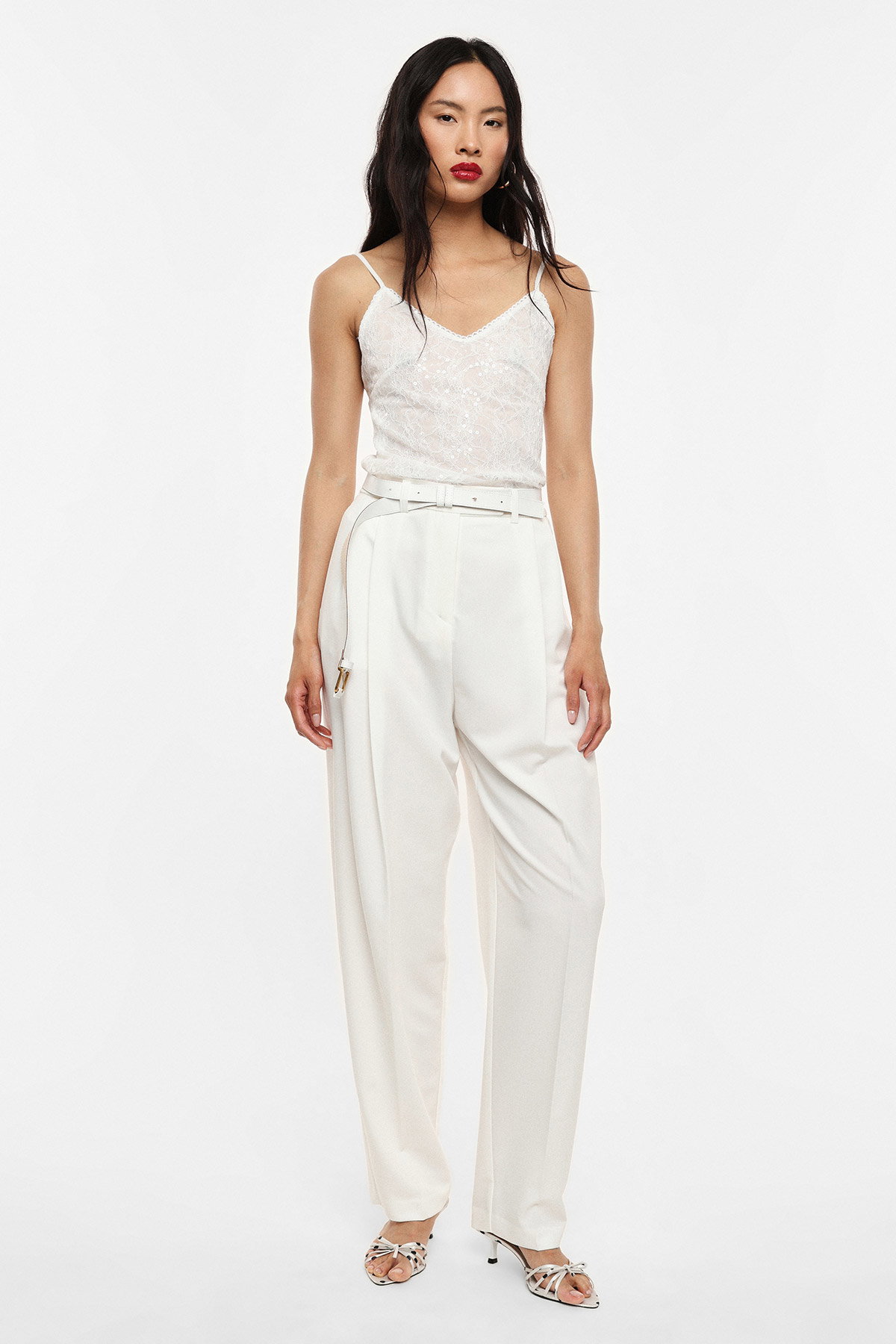Woman model in white sequin camisole and high-waisted wide-leg white trousers with belt and strappy heels on plain background