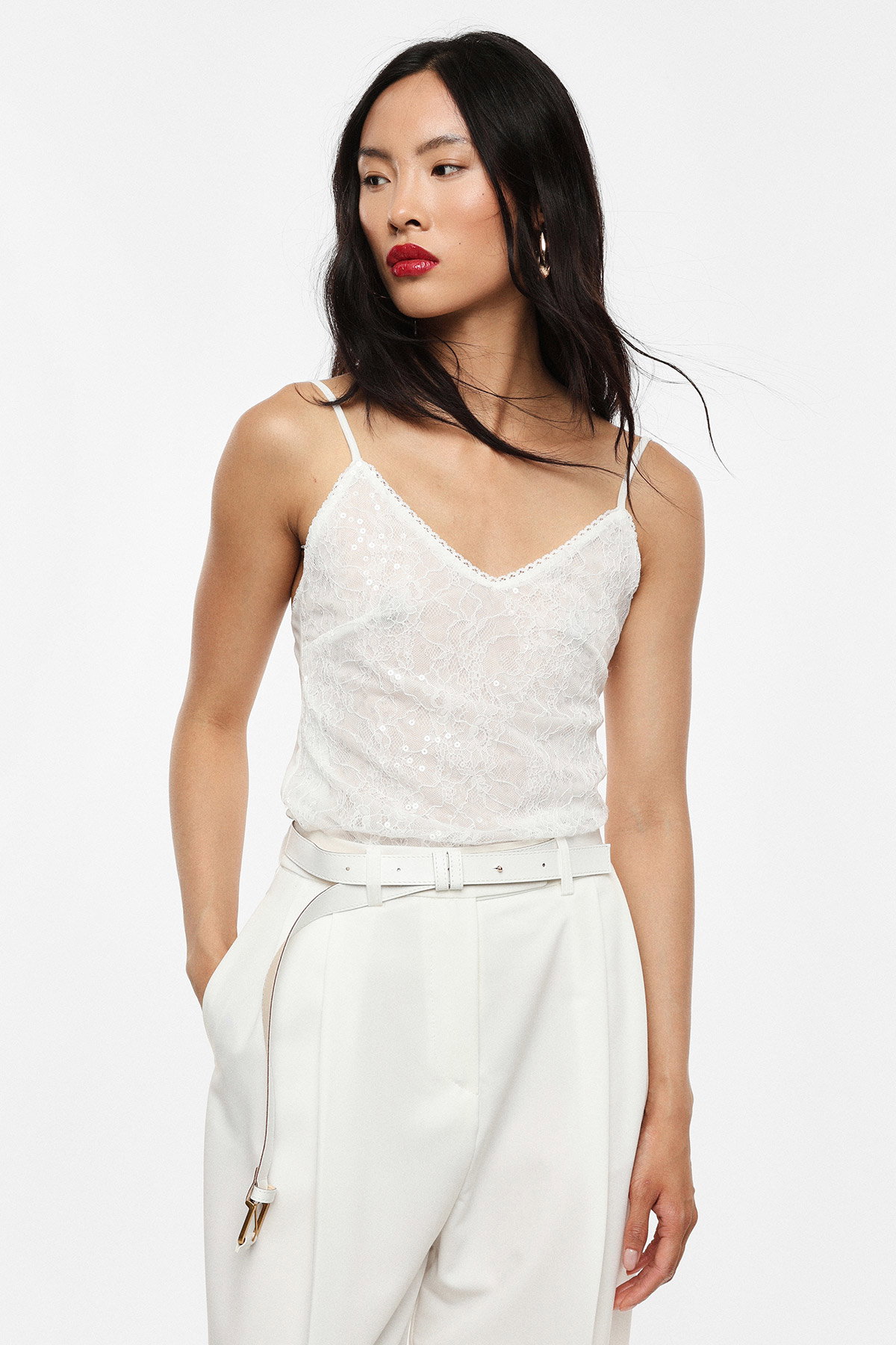 Female model wearing a white lace camisole and high-waisted white trousers with belt, red lipstick and hoop earring, hand in pocket