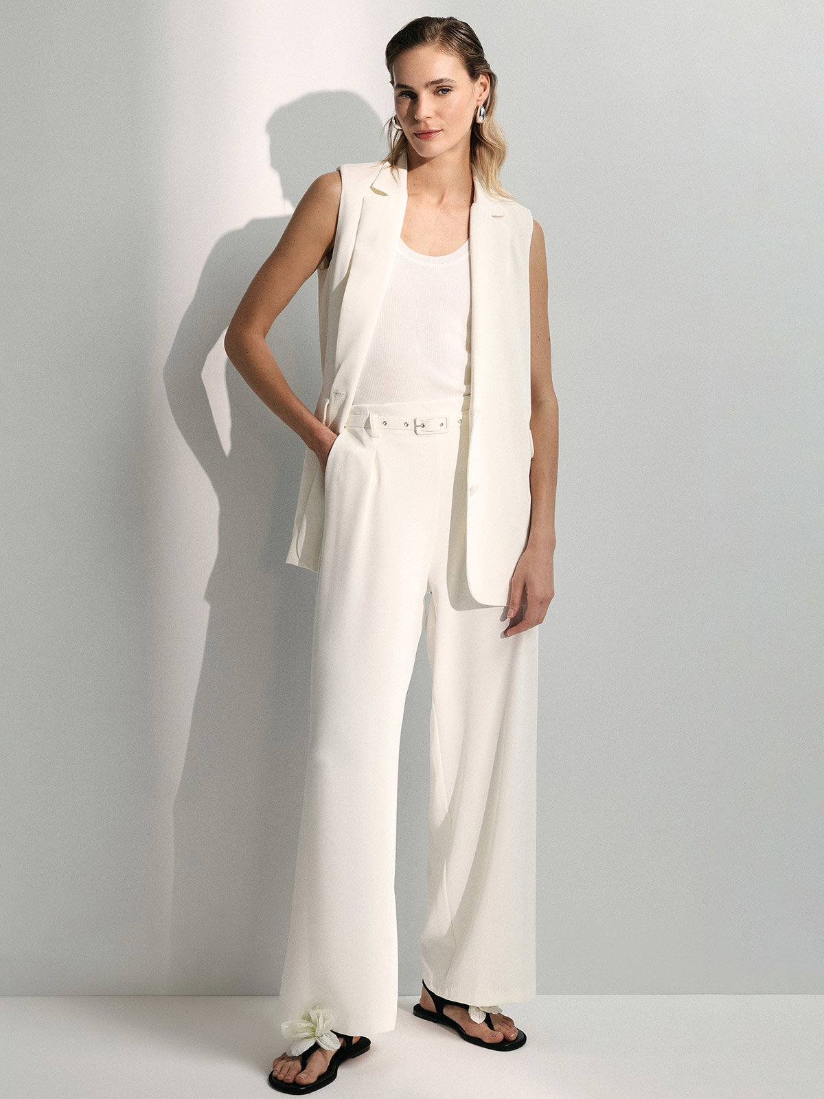 Model in white sleeveless blazer and wide-leg trousers with belt, ribbed tank and black sandals with flower