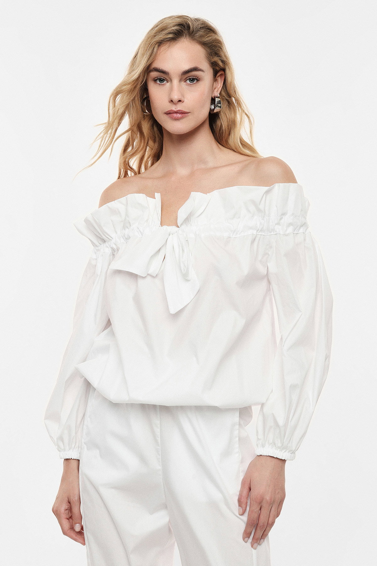 Model wearing white off-shoulder long-sleeve blouse with bow detail and matching white trousers against plain background