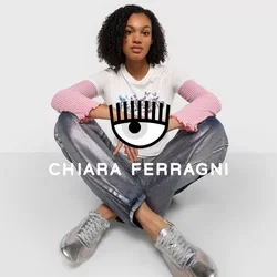 T-shirt bianca con maniche a coste rosa, dotata di una grafica, abbinata a pantaloni metallici argento e sneakers trasparenti, posata su uno sfondo bianco.