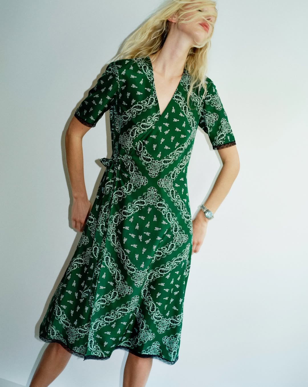 Woman in a green paisley-patterned wrap midi dress with short sleeves and wristwatch, posing against a pale background