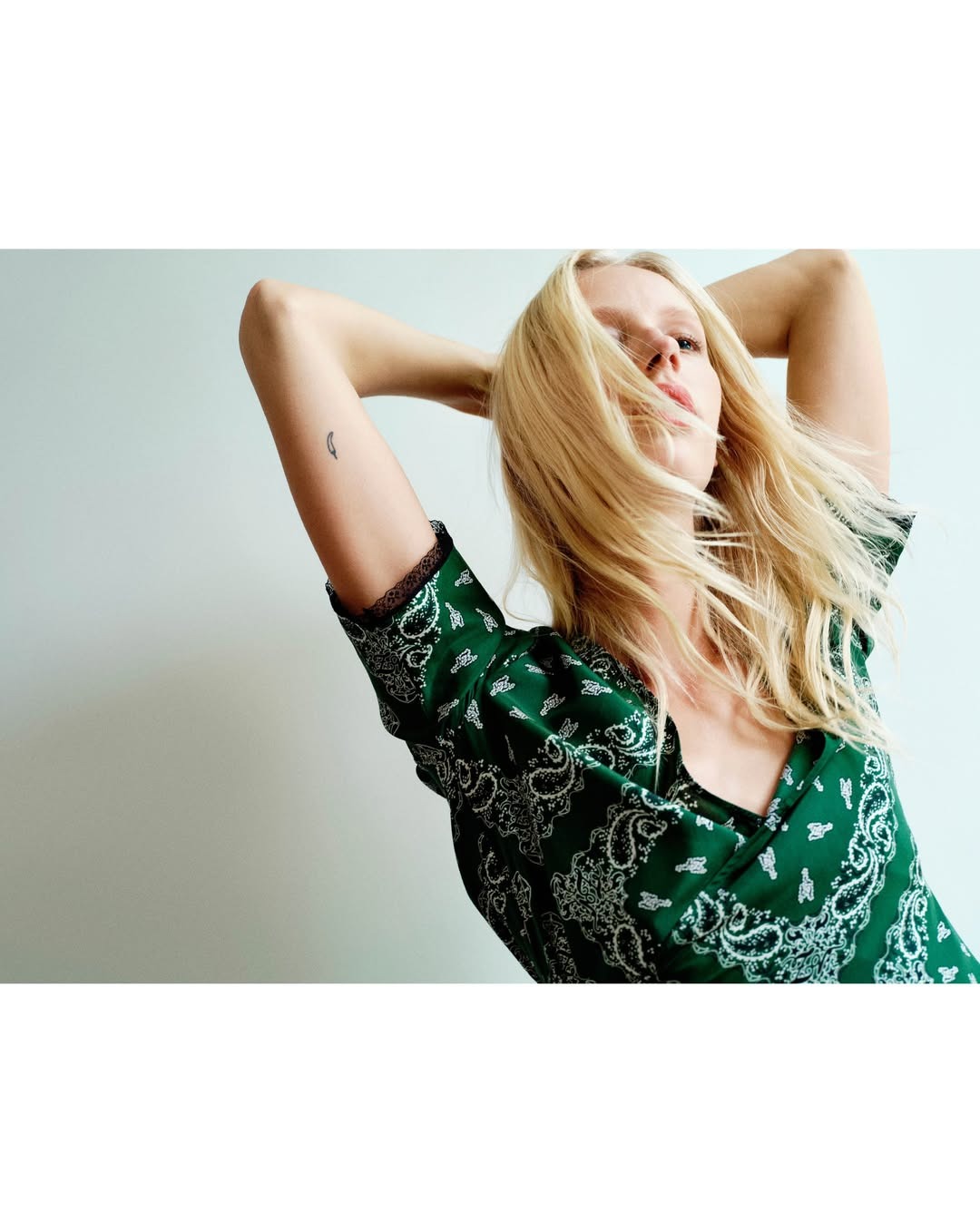 Blonde woman in green paisley dress posing with arms raised, hair partially covering her face — fashion portrait on plain studio background