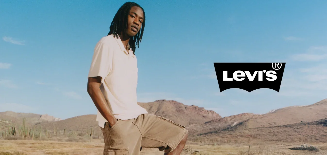 Hombre con rastas que lleva pantalones cortos beige y camisa blanca, equilibra un pie sobre una silla plegable amarilla en un paisaje seco y montañoso bajo un cielo azul.