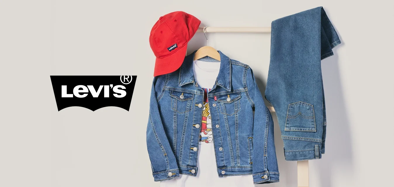 Gorra roja, chaqueta vaquera sobre camiseta blanca y jeans azules doblados colgados en un perchero de madera contra un fondo liso.